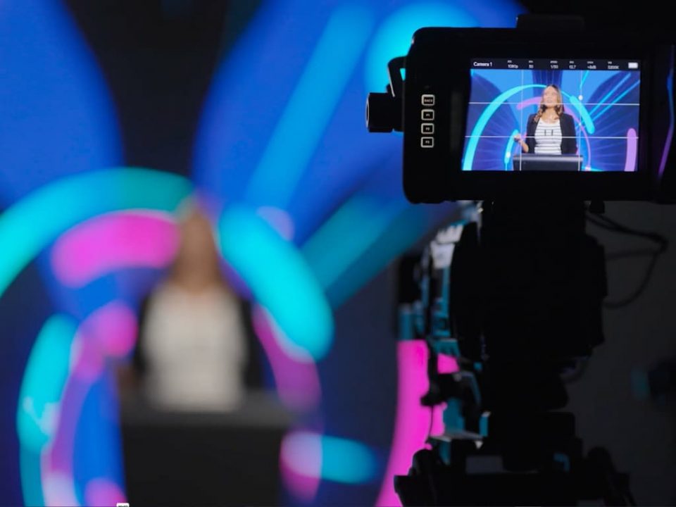 woman standing in front of a LED screen while being recorded by a broadcast camera.