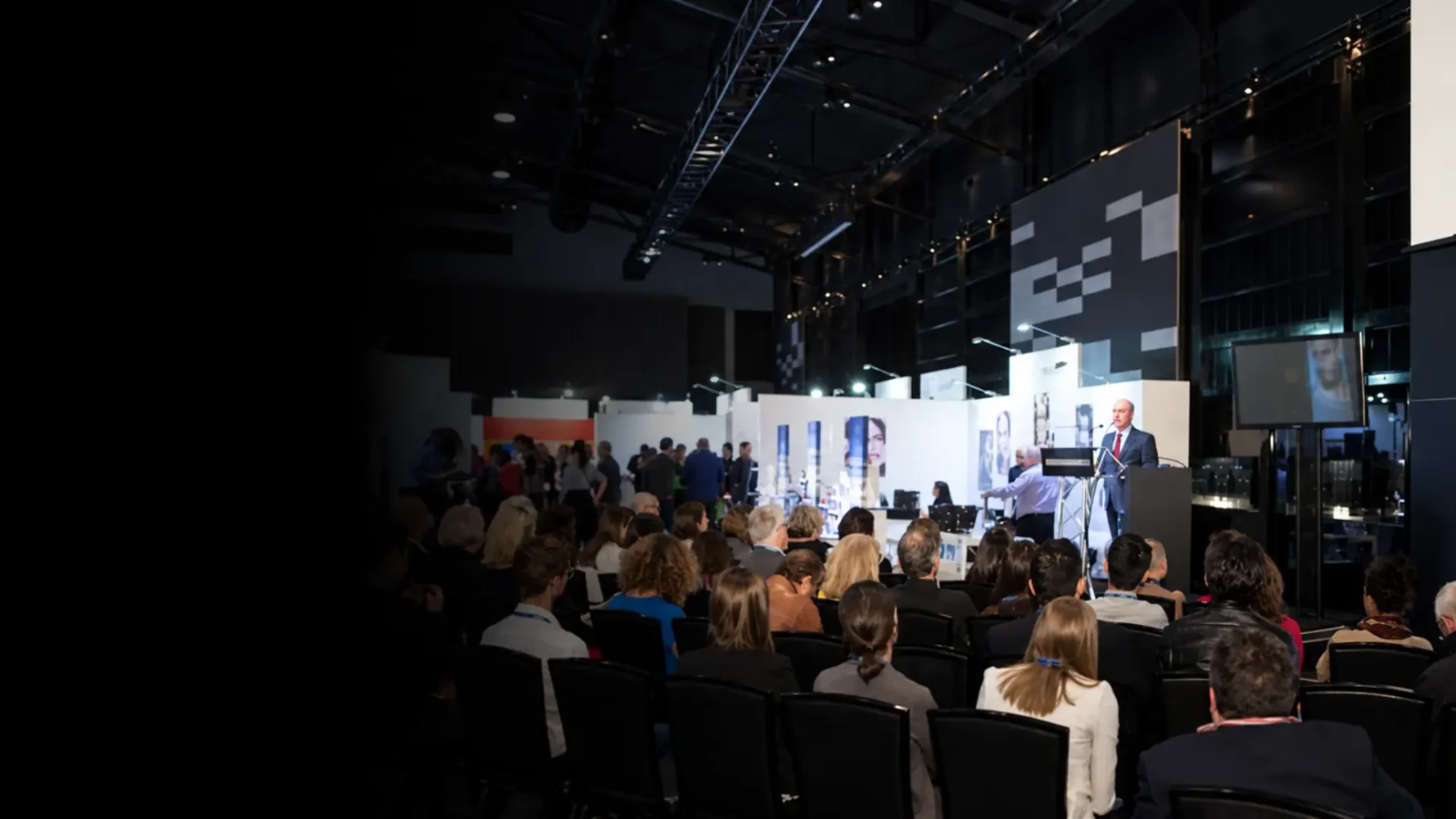Presenter addressing a seated exhibition audience beside a freestanding presentation screen.