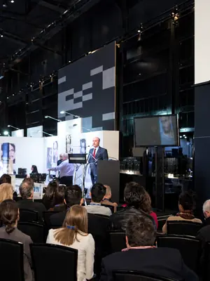 Exhibition speaker at a lectern with a presentation monitor and audience in view.