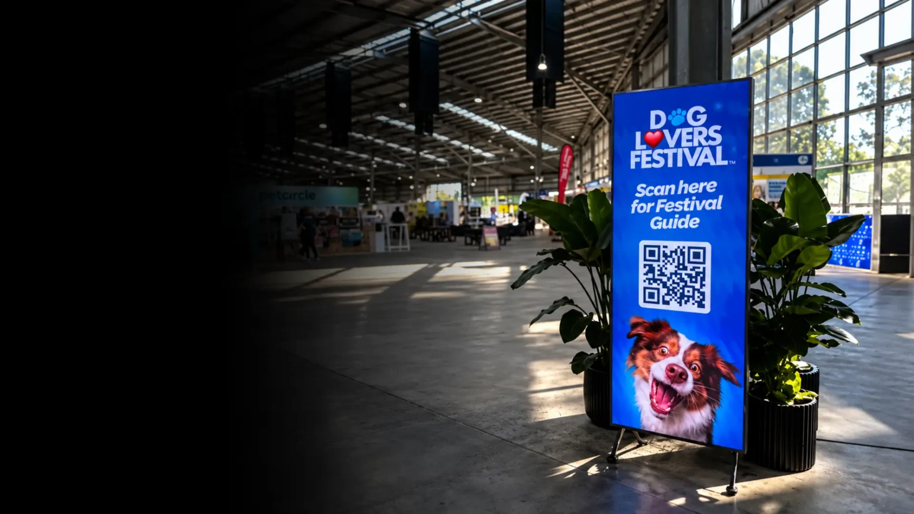 Digital signage hire display showing a Dog Lovers Festival guide beside plants in an exhibition hall.