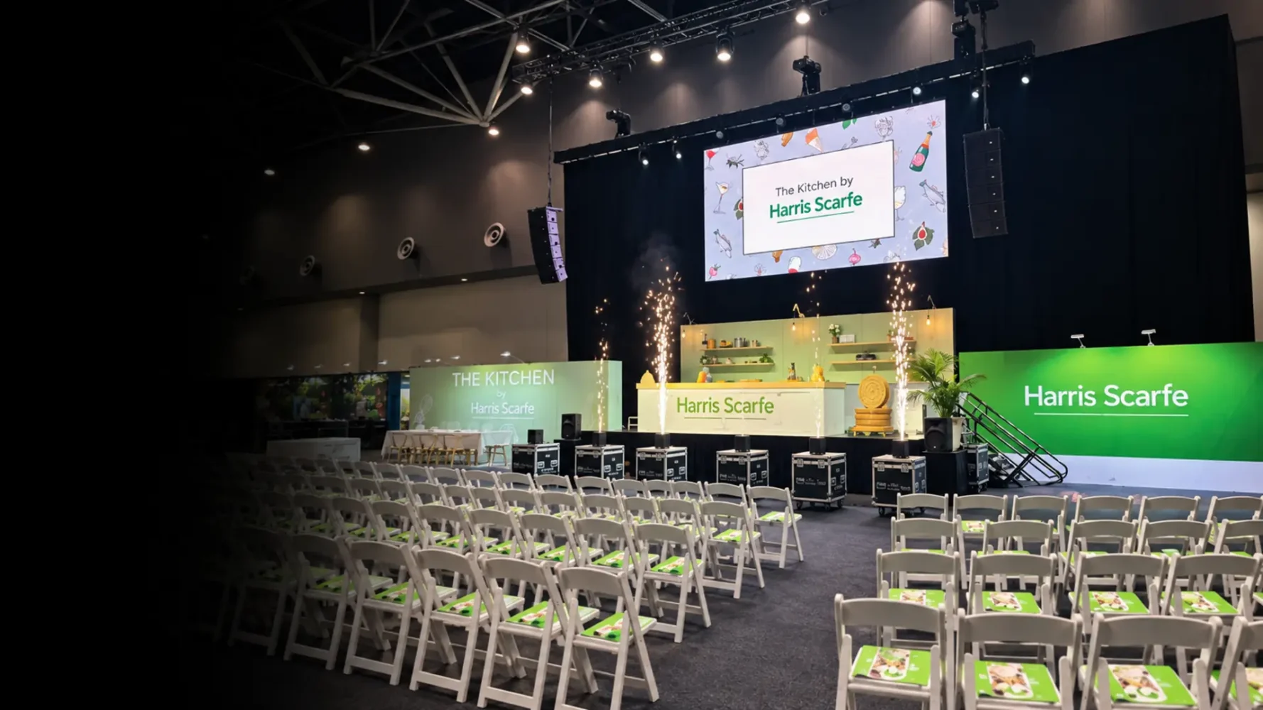 Large digital screen displaying Harris Scarfe branding above a demonstration kitchen and rows of event seating.