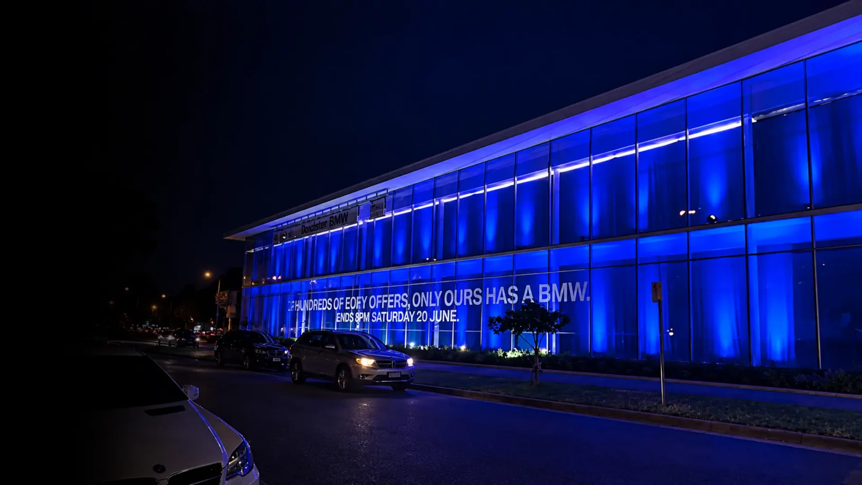Glass-fronted showroom illuminated with blue lighting at night, with vehicles on the road outside.
