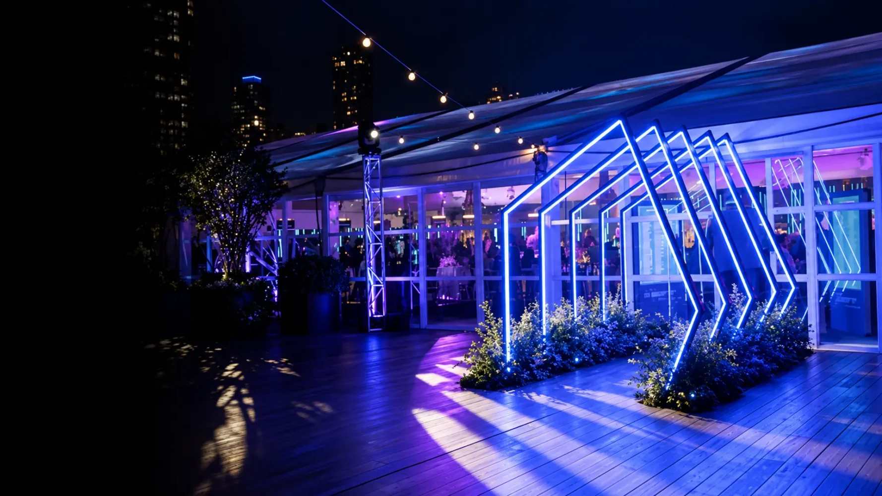 LED entrance hire featuring angular blue-white light frames over a planted walkway outside an event venue.