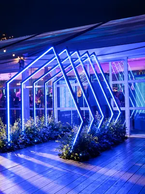 Geometric LED entrance frames illuminating a timber walkway with blue and violet light.