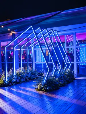 Angular LED frames casting blue and violet light across a planted event entrance and timber walkway.
