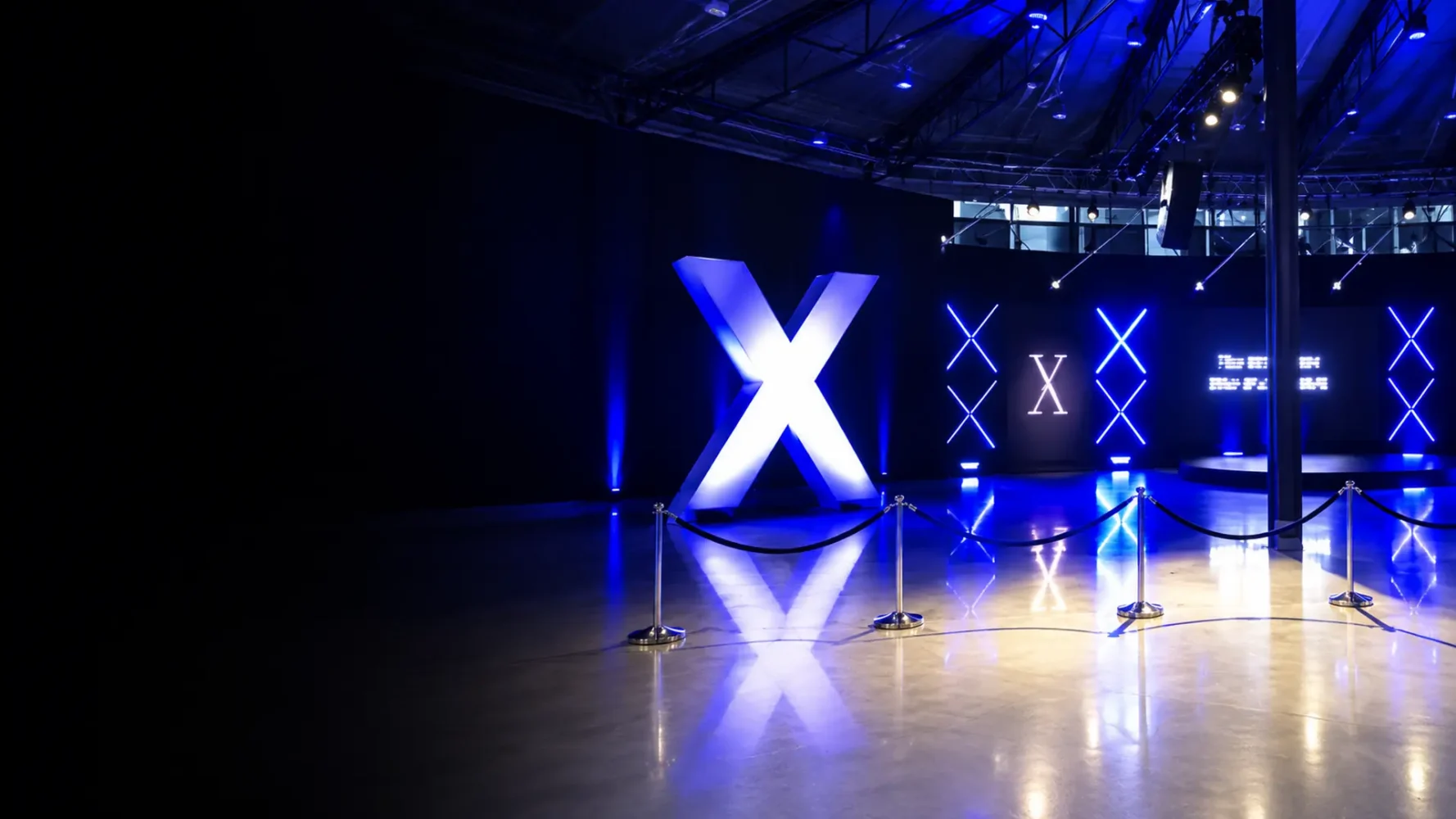 Event lighting display with a large illuminated white X, blue X motifs and reflections across a polished floor.