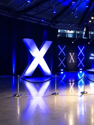 Illuminated X display and blue-lit wall motifs behind rope barriers at an indoor event.