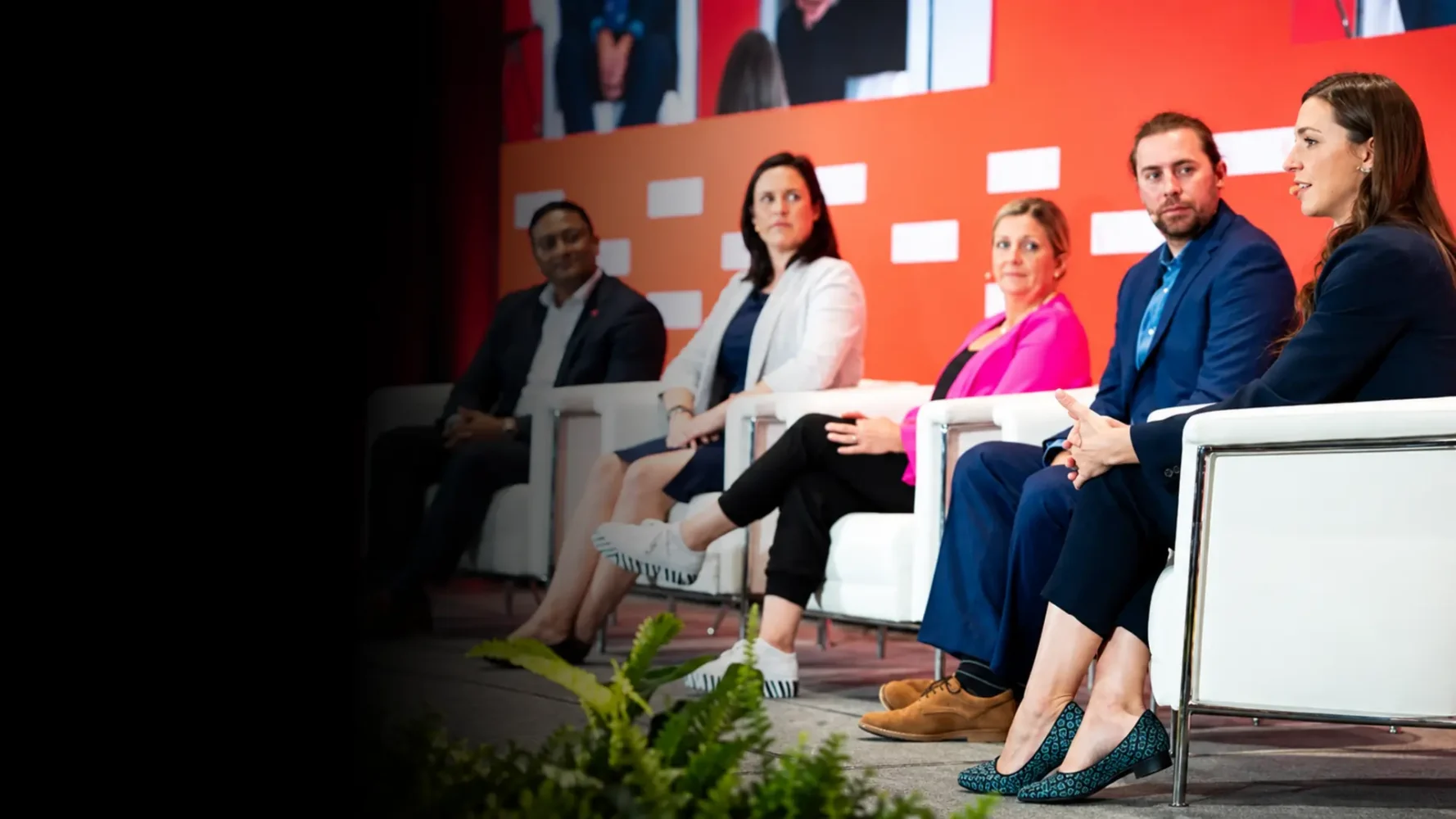 Five conference panellists seated in white armchairs as a speaker addresses the audience.