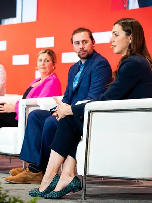 Conference panellist speaking from a white armchair beside fellow presenters.