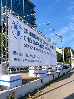 BMW promotional banner supported by a freestanding truss structure beside a footpath.