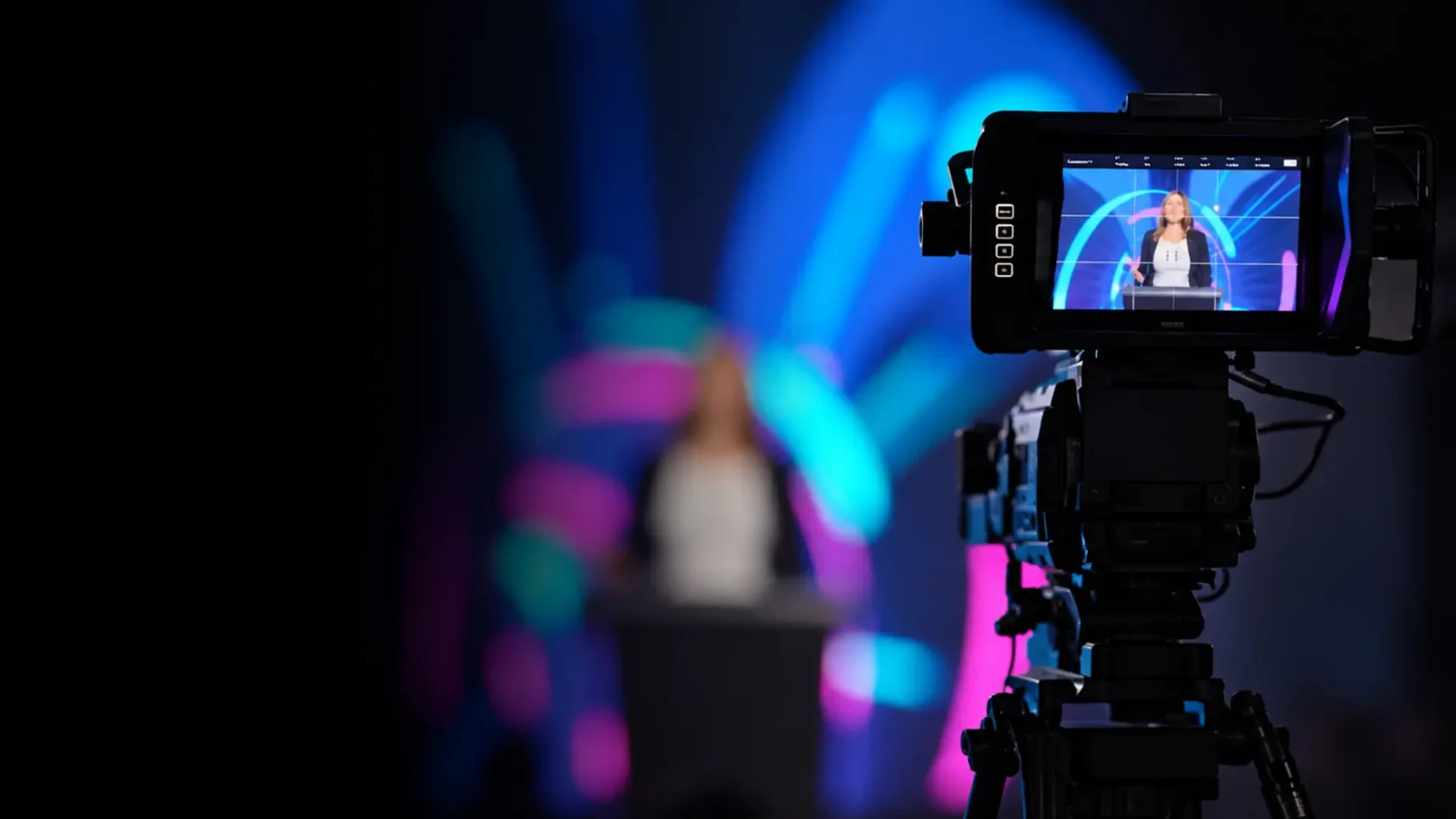 Professional video camera filming a presenter at a lectern against blue and magenta event lighting.